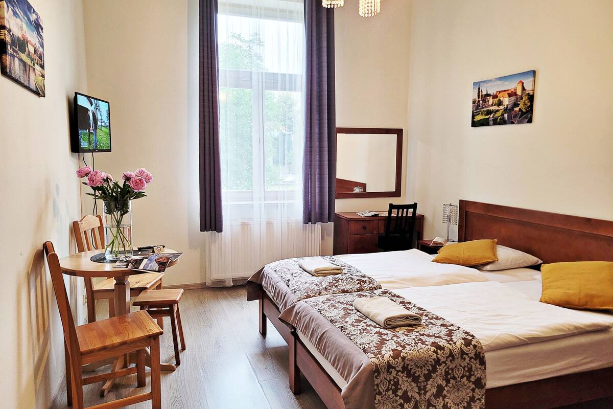 Una camera d'albergo con due letti, una scrivania, una sedia, un tavolo da pranzo e una finestra con tende.