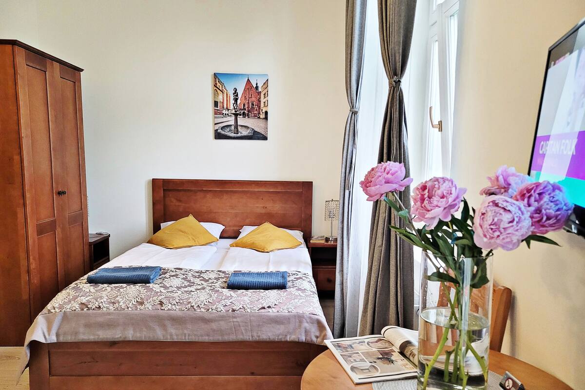 Un letto rifatto con cura, con una testiera in legno, due cuscini e una coperta con motivo in pizzo. Su un comodino in legno accanto al letto, un vaso con fiori.