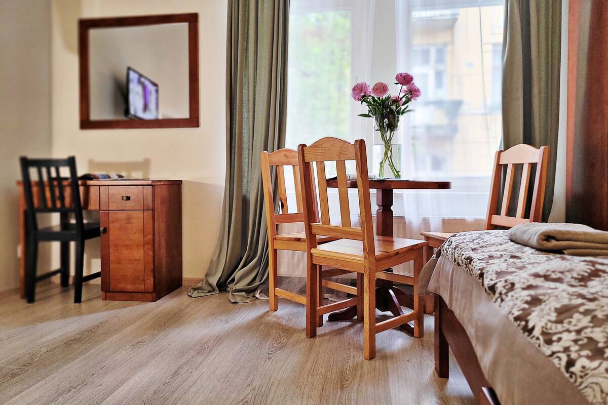 Una camera d'albergo con una scrivania in legno, una sedia, un letto con coperta a fantasia, un piccolo armadietto e una finestra con tende.