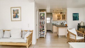 A cozy living room with a sofa and a wicker chair, a bookshelf filled with books, and a kitchen area with cabinets and a sink. - 10 Person Holiday Home in Graested (Graested)