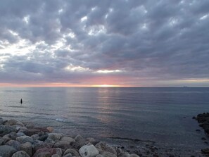 A rocky shoreline with a person standing in the water, a sunset in the background, and a ship on the horizon. - 10 Person Holiday Home in Graested (Graested)