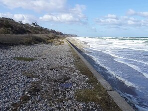 A rocky beach with waves crashing onto the shore, a concrete path along the water, and a hilly landscape in the background. - 10 Person Holiday Home in Graested (Graested)