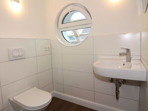 A small, clean bathroom with white tiles, a round window, a white sink, and a toilet. - Ferienhaus in Wendorf in Meeresnhhe (Kuhlen-Wendorf)