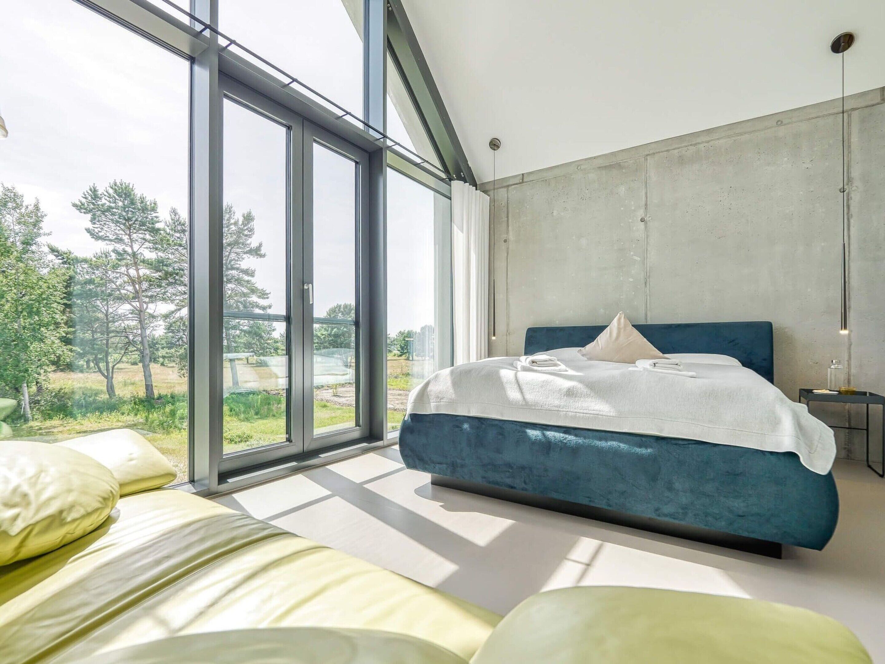 A modern bedroom with a large window, a bed with blue headboard, and a view of greenery outside.