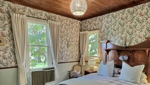 Une chambre avec un plafond en bois, un papier peint à motifs floraux, un lit recouvert d’une courtepointe à carreaux, un banc et une fenêtre avec des rideaux.