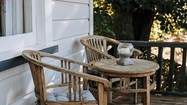 Une terrasse en bois avec deux chaises et une table, un bâtiment blanc en arrière-plan et un arbre sur la droite.