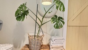 Une malle en osier contenant un pot de monstera, une lampe suspendue et des serviettes pliées.