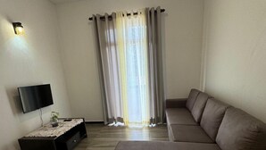 A living room with a sectional sofa, a flat-screen TV, a small table with a vase, and a window with curtains. - Elegant family friendly villa close to airport & beach (Pamunugama)