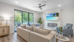A modern living room with a beige sectional sofa, a fireplace, a ceiling fan, and a large window overlooking greenery.