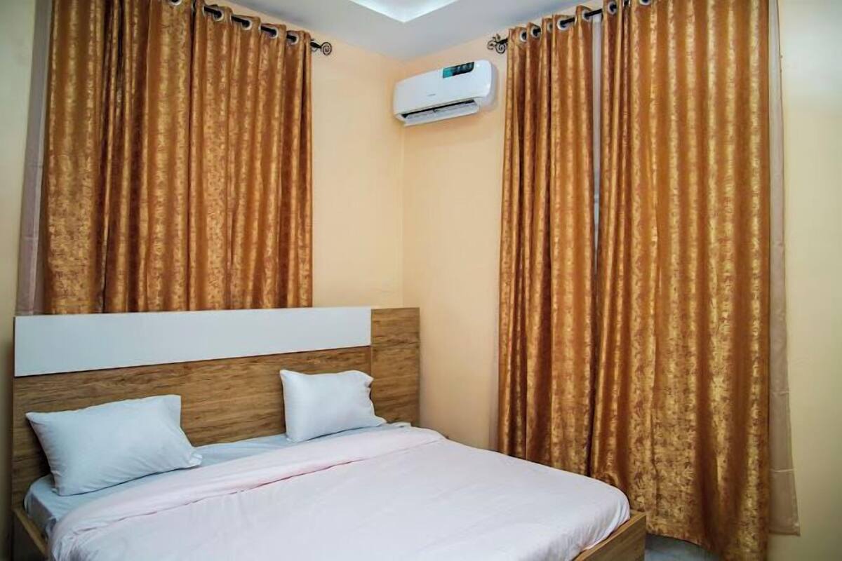 Une chambre d’hôtel avec un lit, deux oreillers, une tête de lit et une fenêtre avec des rideaux.