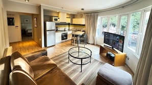 Una sala moderna con sofá, mesa de centro y televisor. Cuenta con una cocina equipada con refrigerador, microondas y estufa. La habitación tiene ventanas amplias con cortinas.