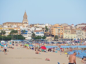 Ein überfüllter Strand mit Menschen, die sonnenbaden und schwimmen, und ein historisches Gebäude mit einem Glockenturm im Hintergrund.