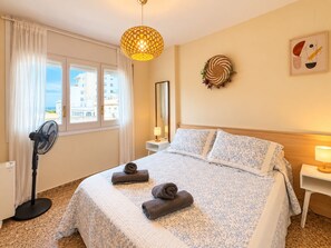 A hotel room with a bed, a standing fan, a window with curtains, a wall-mounted mirror, and a decorative wreath.
