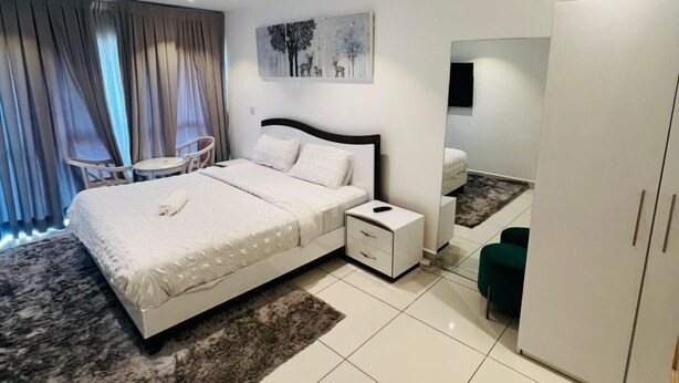 A modern bedroom with a large bed, a bedside table, a wardrobe, a small desk, and a chair. - Prime properties executive two bedroom  (Accra)