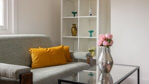 Una sala de estar moderna con un sofá gris, un cojín amarillo, una mesa de centro de vidrio y un estante blanco con objetos decorativos.