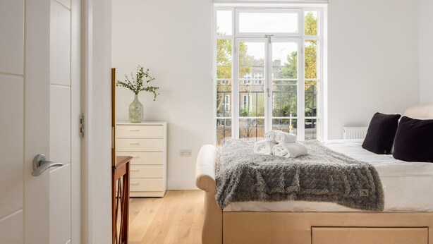 A bedroom with a large window, a bed with a grey blanket, a vase with plants, and a wooden nightstand. - Picktheplace Kensington Lux Apartments (London)