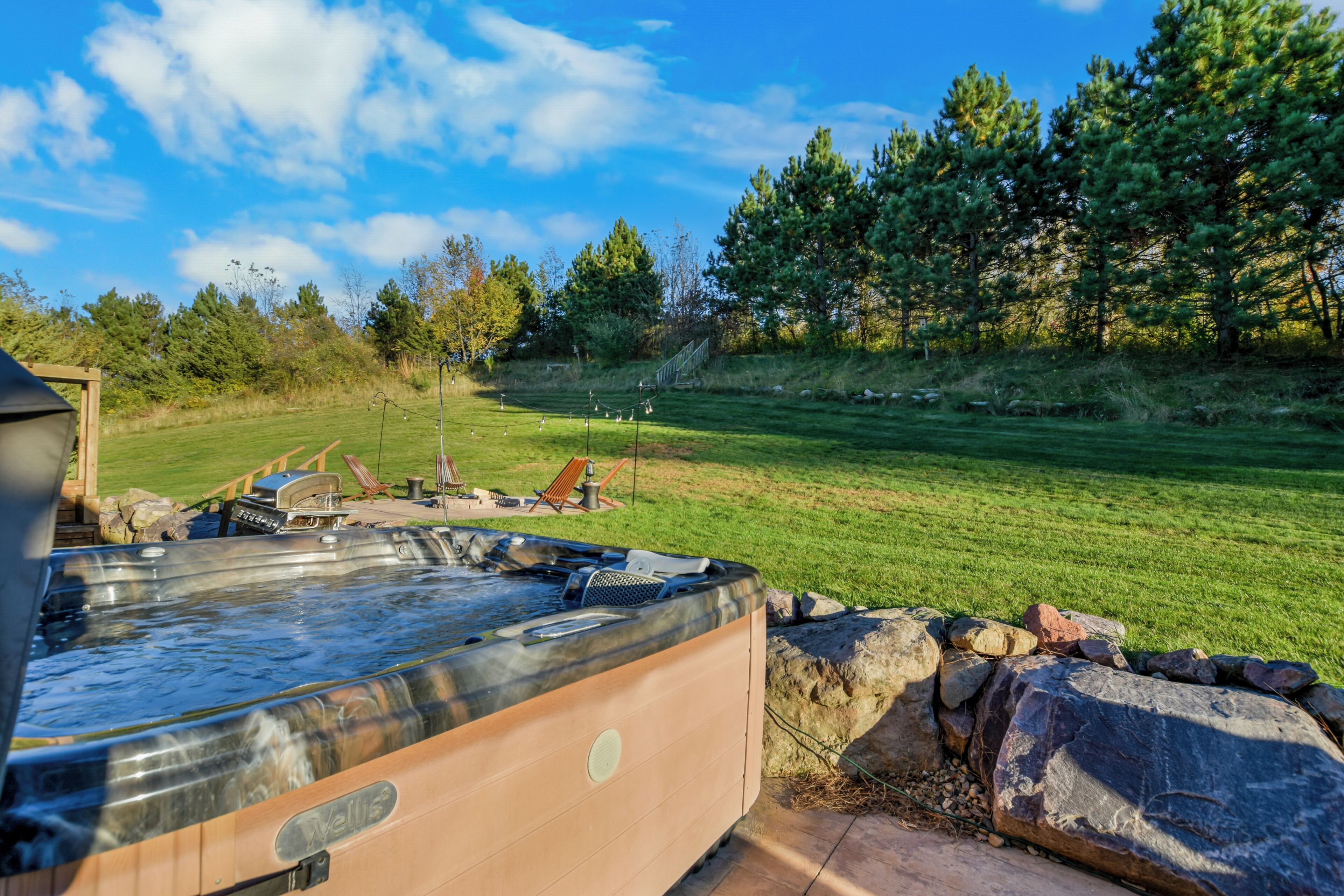 A hot tub in a backyard with a grassy lawn, trees, and a wooden deck.