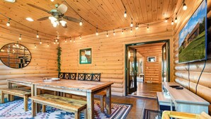 Interior d'una cabana de fusta amb una taula de dinar de fusta, tamborets, un ventilador de sostre, llums de garlandes, un mirall i una televisió penjada a la paret.