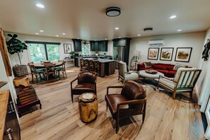 A spacious living room with a wooden floor, a dining area with a large table, and a kitchen with dark cabinets and a refrigerator.