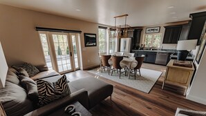 Una sala de estar moderna con sofá, reposapiés y mesa de centro, un comedor con sillas y una mesa, y una cocina con armarios oscuros y un refrigerador.