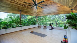 Une pièce spacieuse avec un plafond en bois, de grandes fenêtres, un ventilateur de plafond, du matériel d’exercice et un tapis de yoga.