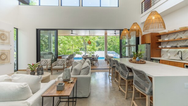 Une cuisine moderne dotée d’un grand îlot central, d’un coin repas avec des chaises et offrant une vue sur la verdure environnante.
