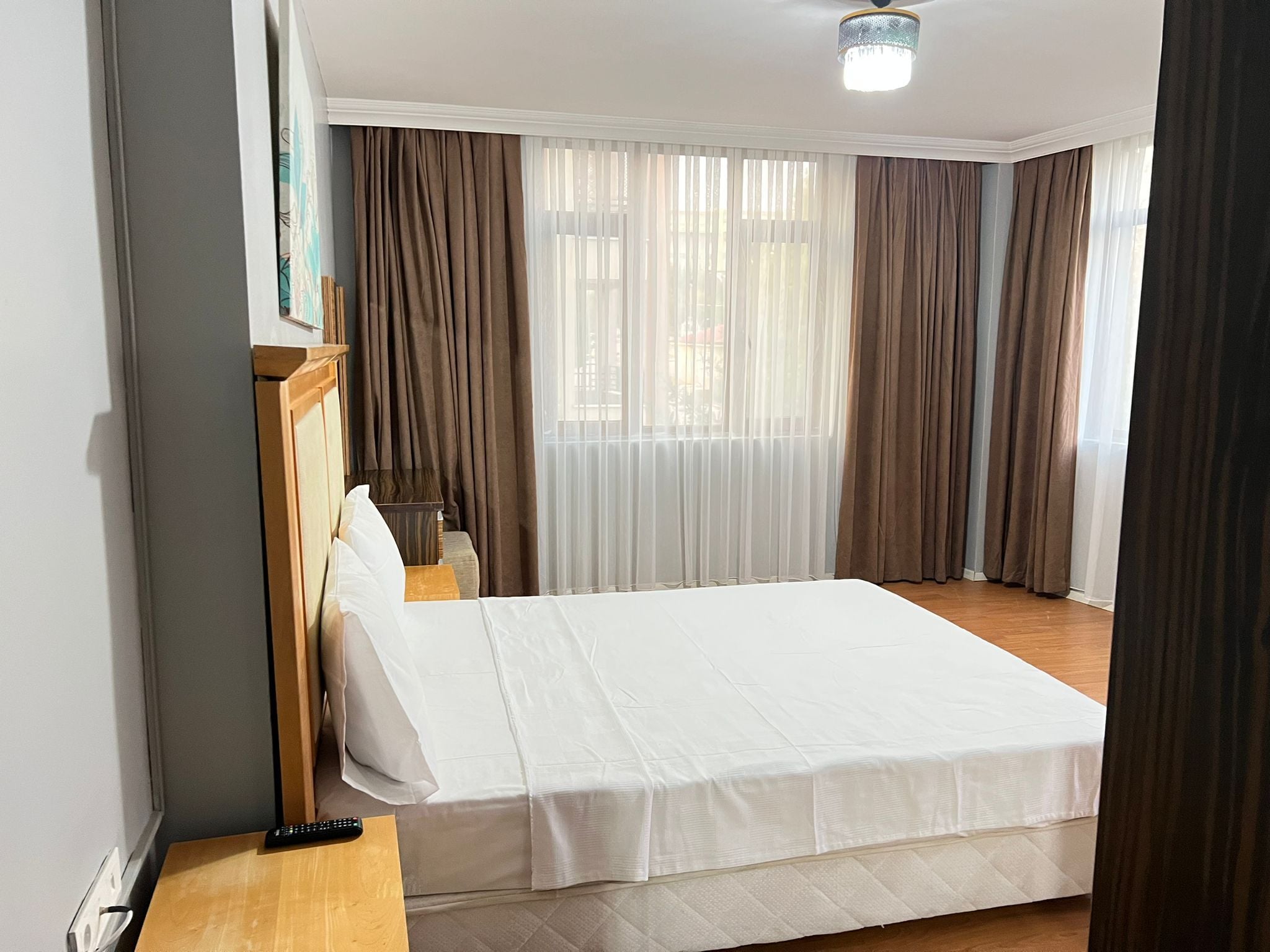 A hotel room with a bed, a wooden nightstand, a remote control, and a window with curtains.