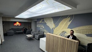 A modern reception area with a high ceiling, a large wall mural, and a flat-screen TV displaying a fireplace. - City Blue Hotel (Elazig)