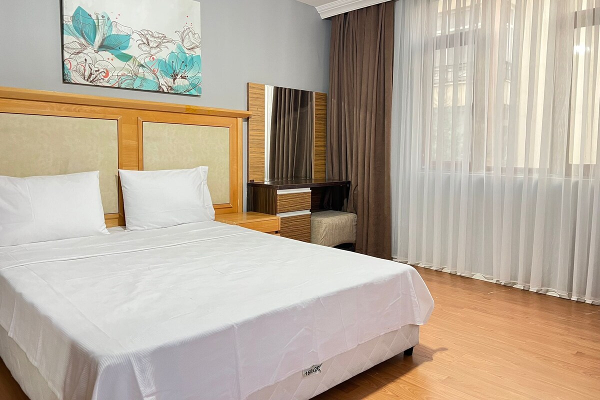 A hotel room with a large bed, a wooden headboard, a flat-screen TV, a small armchair, and a large window with sheer curtains.