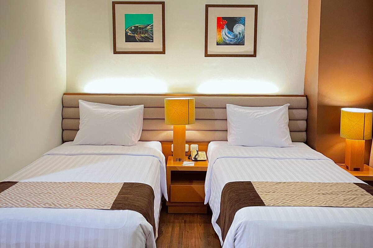 A hotel room with two beds, a nightstand with a lamp, and two framed pictures on the wall.