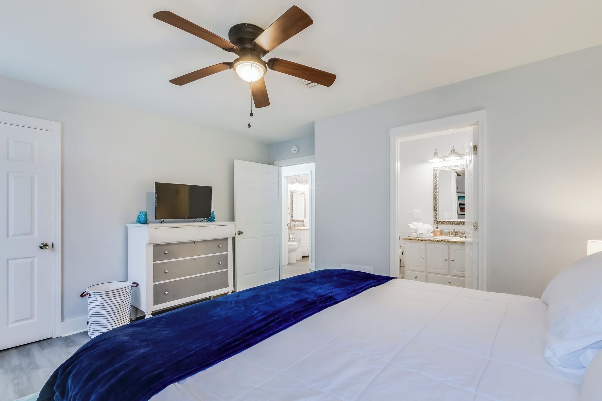 Camera da letto con un letto, una ventola a soffitto, una televisione su un comò e un bagno visibile attraverso una porta aperta.