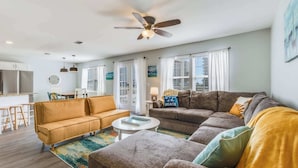 A spacious living room with a sofa, ottoman, and a coffee table. There is a dining area with a table and chairs, and a kitchen with a refrigerator. - Sel De Vie A 4 Bedrooms Sleeps 10 (Gulf Shores)