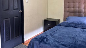 A bedroom with a dark wooden door, a small bedside table, a bed with a blue bedspread, a wall-mounted air conditioner, and a framed picture on the wall. - HOTEL KOREA GARDEN (Apodaca)