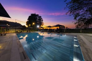 A swimming pool with a tiled deck, surrounded by umbrellas and benches, set against a sunset sky.