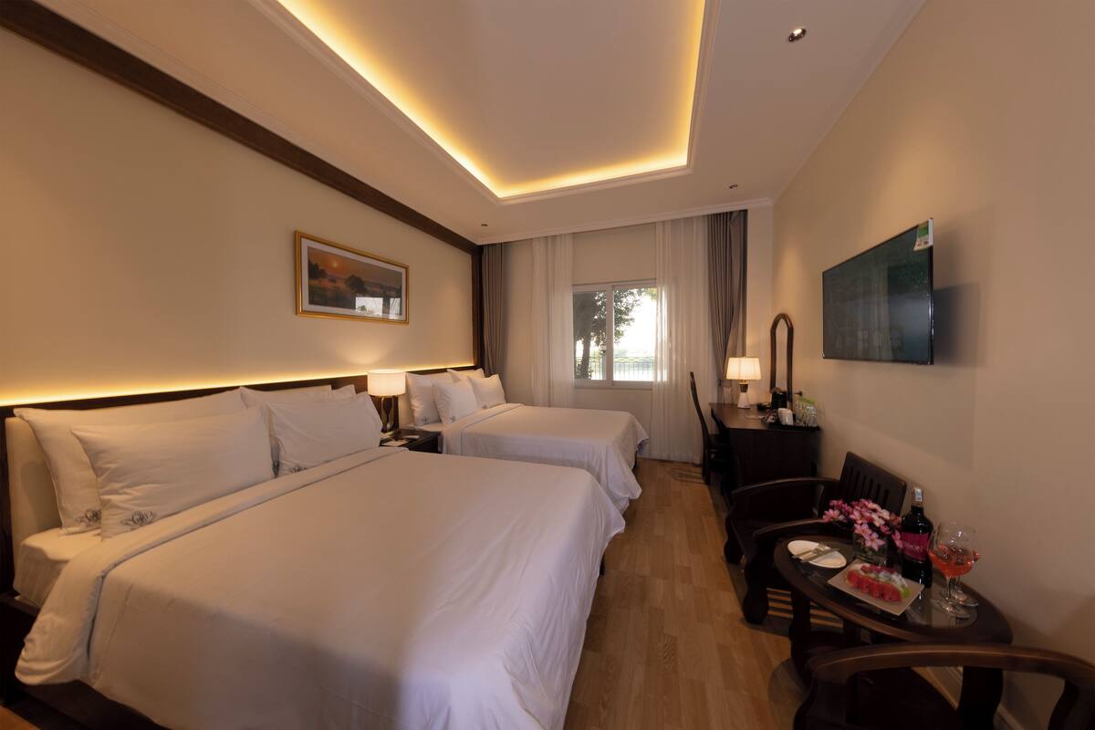 A hotel room with two beds, a dining table with chairs, a TV, and a window with curtains.