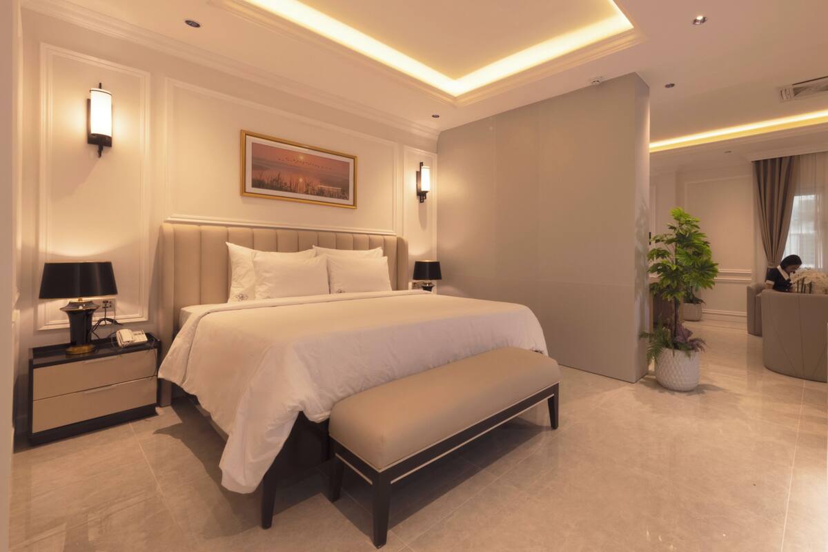 A modern hotel bedroom with a large bed, bedside tables, a bench, a potted plant, and a painting on the wall.