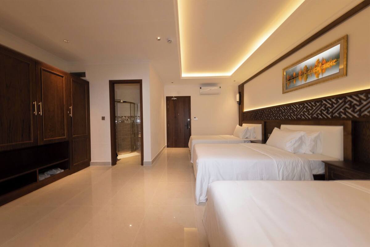 A hotel room with three beds, a wardrobe, a shower area, and a painting on the wall.
