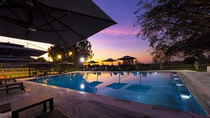 Une piscine entourée d’arbres, avec des chaises longues et des parasols, et un coucher de soleil en arrière-plan.