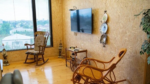 A cozy room with a large window, a wooden rocking chair, a small table, and a flat-screen TV mounted on the wall. - Hotel bani (Kutaisi)