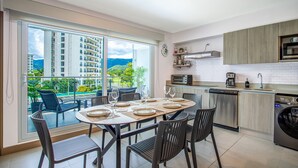 A modern kitchen with a view of a cityscape, a dining area with a table set for four, and a balcony with chairs. - Casago | Beach & Marine Design Condo in Jacó (Jaco)