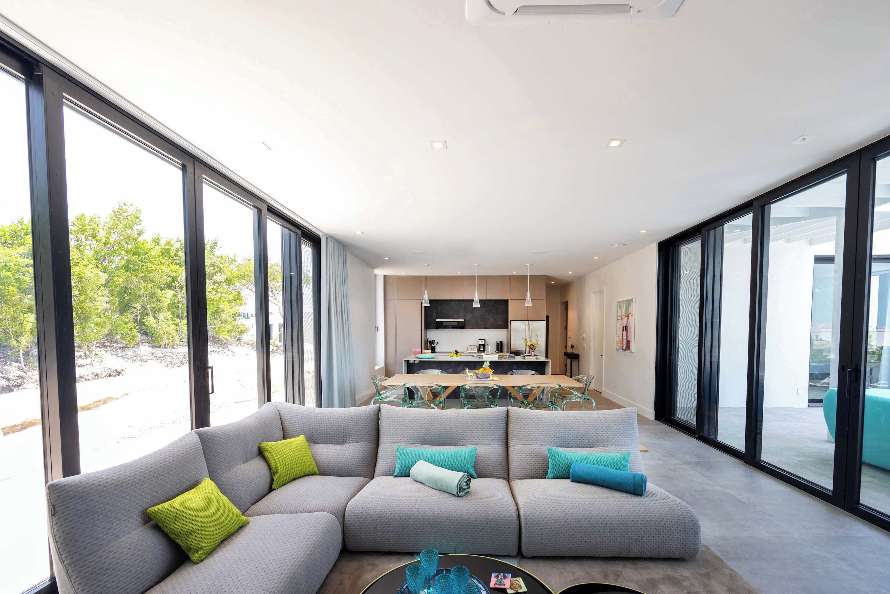 A modern living room with a sectional sofa, a dining area with a glass table, and large windows offering a view of the outdoors.