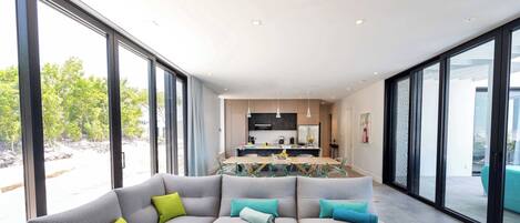 A modern living room with a sectional sofa, a dining area with a glass table, and large windows offering a view of the outdoors.