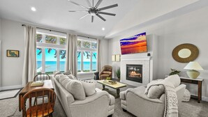 A living room with a fireplace, a ceiling fan, a sofa, a coffee table, and a view of the ocean through large windows. - Spacious 4-bedroom lakefront house with WiFi, AC in tranquil Houghton Lake (Houghton Lake)