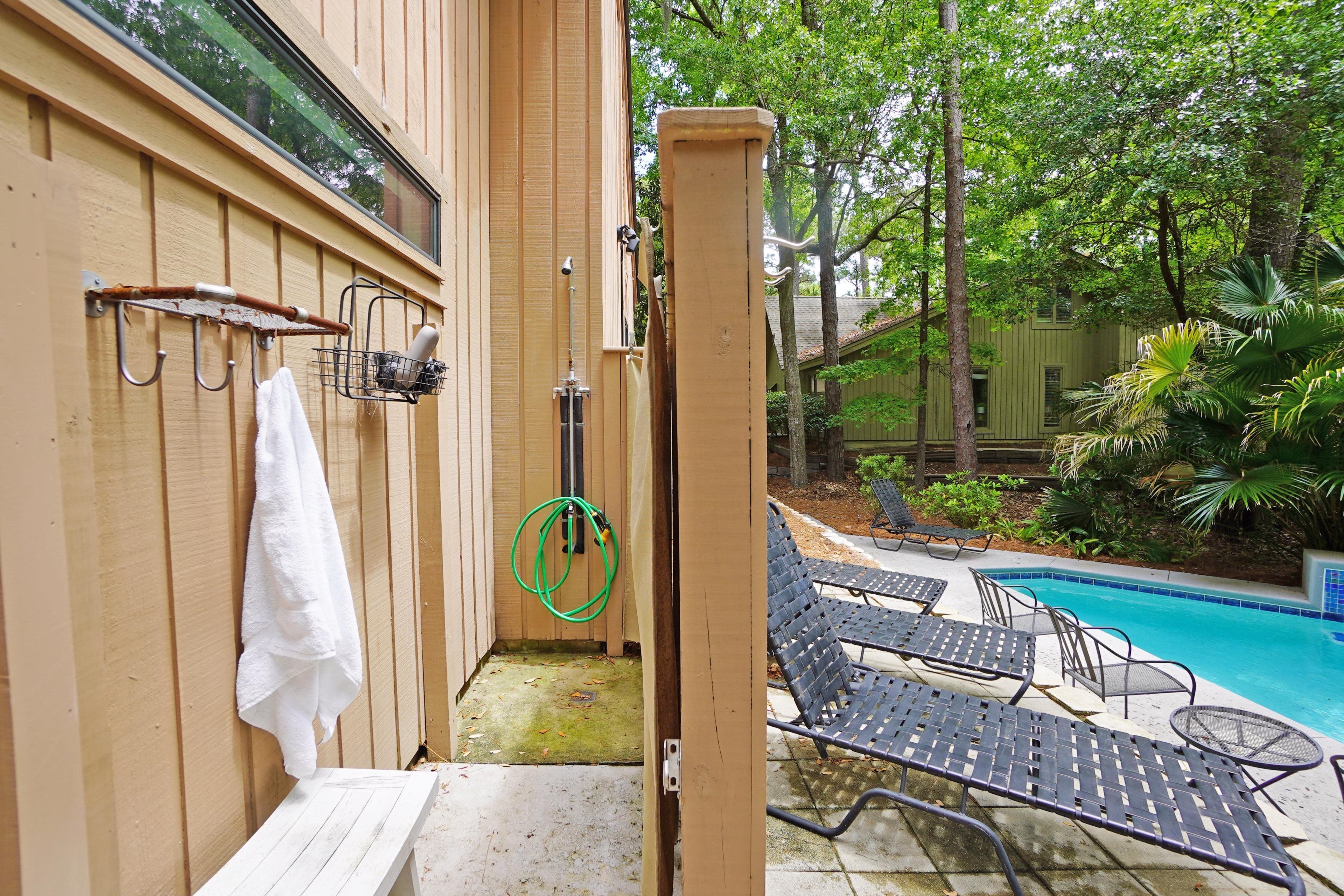 Un mur extérieur en bois, muni d’un porte-serviettes, d’un tuyau d’arrosage et d’une petite porte donnant accès à un espace piscine équipé de chaises longues et d’une piscine.