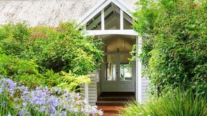 A quaint, white cottage with a steeply pitched roof, surrounded by lush greenery and flowering plants.