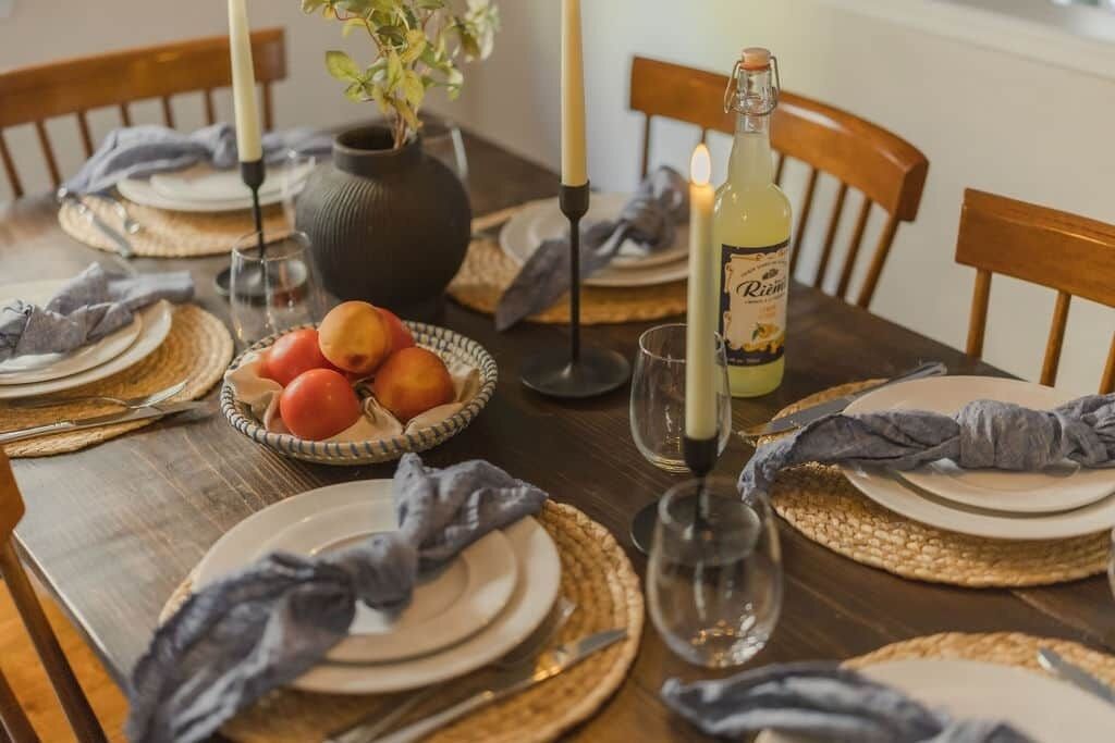 A dining table set with plates, cutlery, a candle, a bottle of wine, and a bowl of fruit.