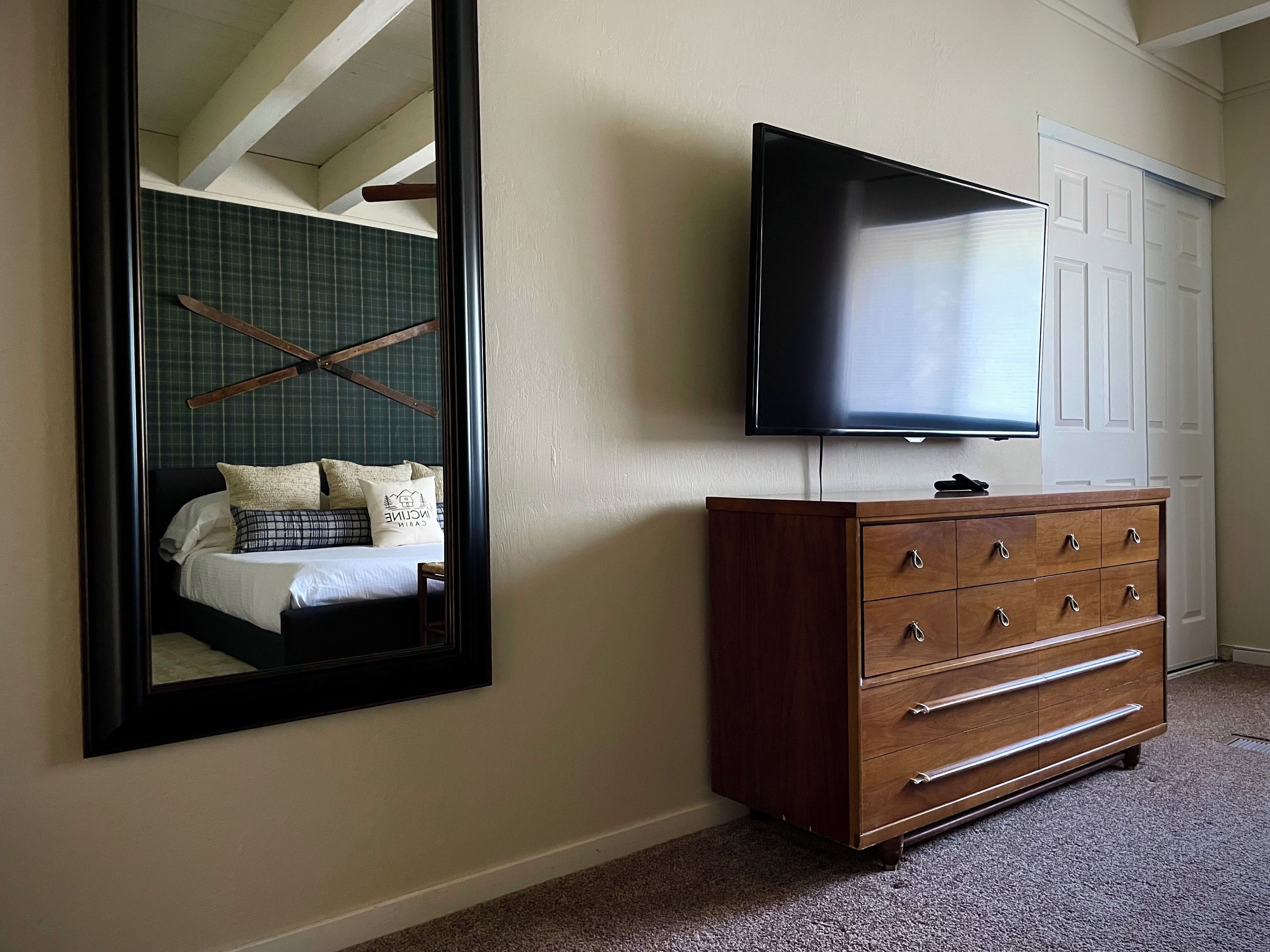 A bedroom with a large mirror, a wooden dresser, and a flat-screen TV mounted on the wall.