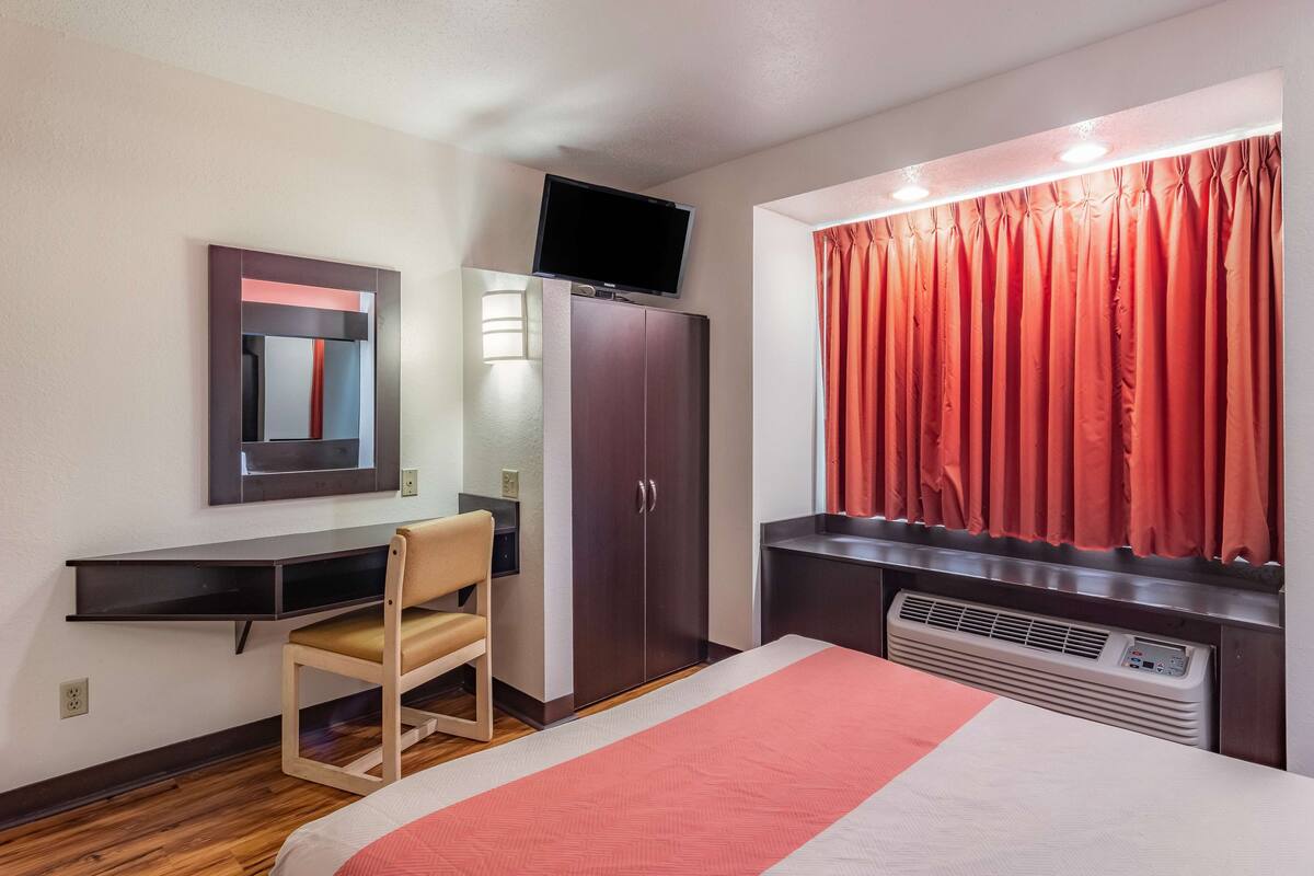 Une chambre d’hôtel comprenant un lit, un bureau avec une chaise, une armoire, une télévision, une fenêtre avec des rideaux et un radiateur.