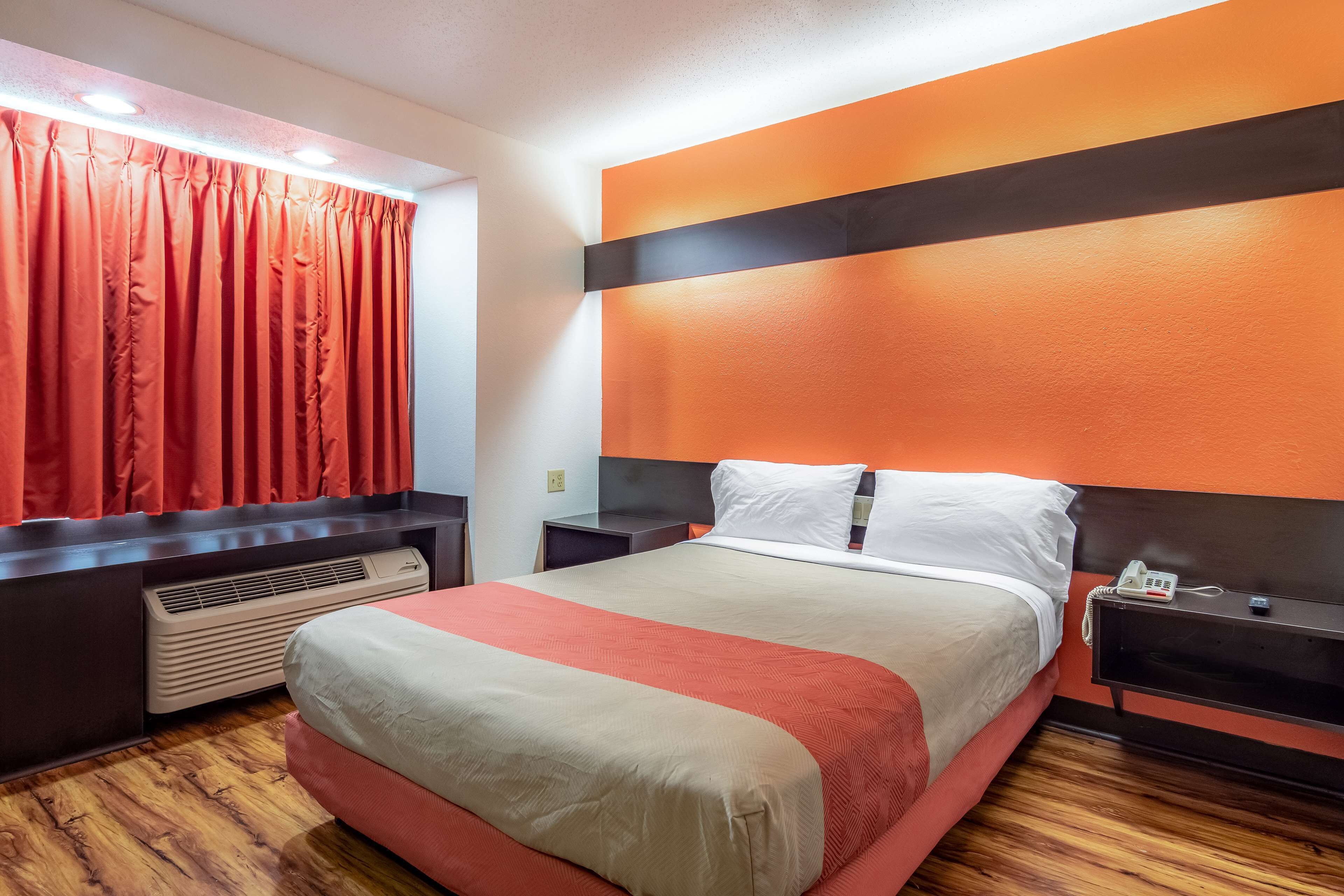 A hotel room with a bed, a nightstand, a heater, and a window with red curtains.