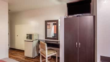 Una habitación pequeña con suelo de madera, un escritorio con silla, un armario, un televisor sobre el escritorio y un refrigerador pequeño.